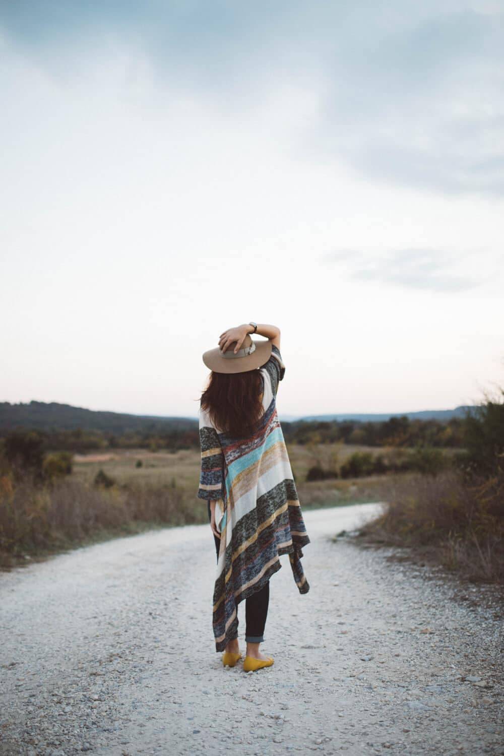 brooke-cagle-ICFPv3xJY90-unsplash | Peru Travel Blog Person standing on a rural gravel road, seen from behind, wearing a multi‑colored striped poncho and a wide‑brim hat.