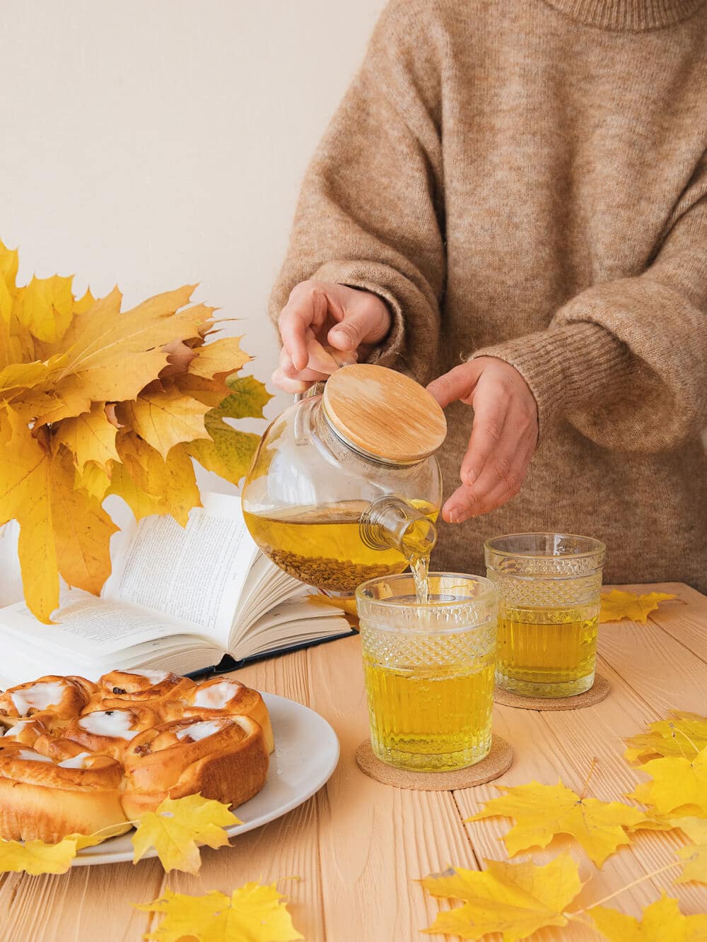 A person in a beige sweater pours tea from a glass teapot into two textured glasses on a wooden table, with autumn leaves and an open book nearby.