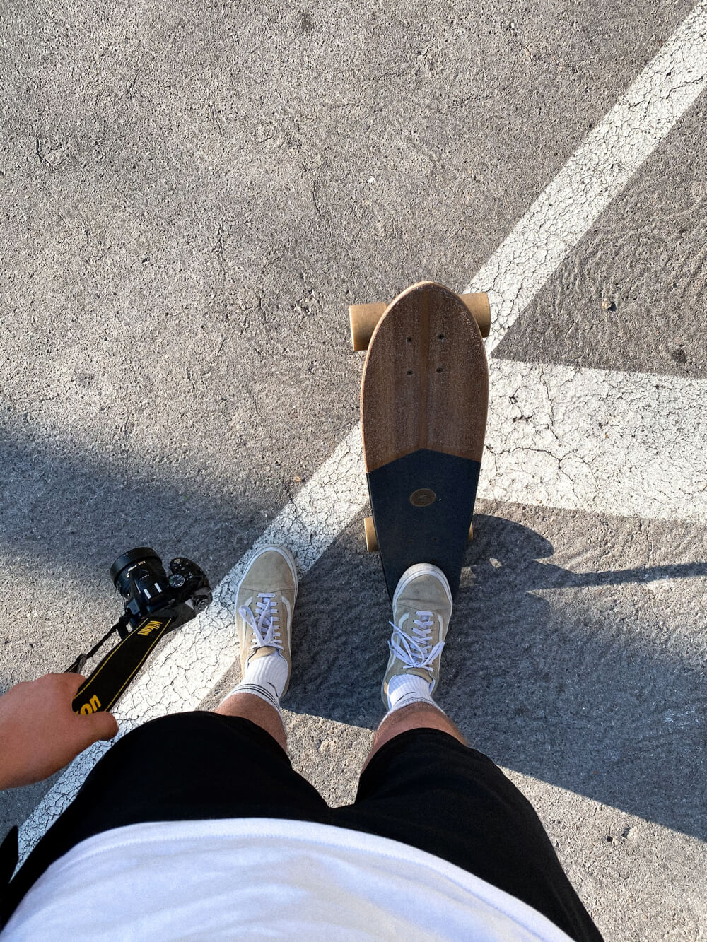 Overhead view of a person wearing white sneakers and black shorts standing with a longboard on cracked pavement near a white line, holding a camera in one hand.