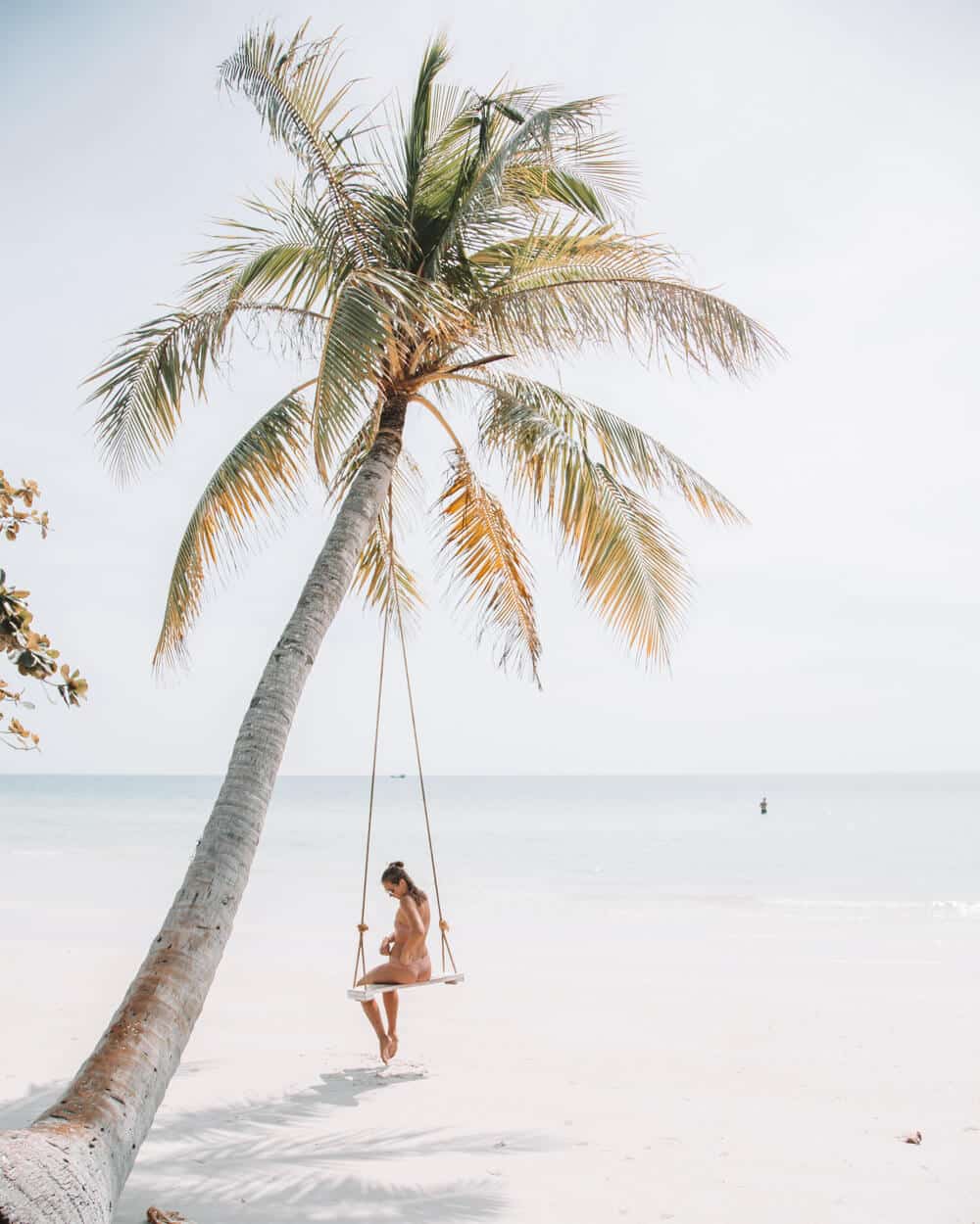 ryan-christodoulou-LDrSJ3cjauY-unsplash | Peru Travel Blog Person lounging on a wooden swing hanging from a leaning palm tree on a sunny, white-sand beach by the calm sea.