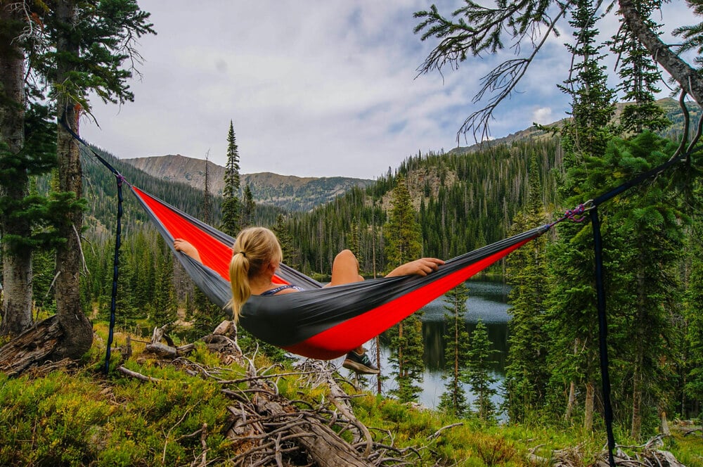 zach-betten-KYTT8L5JLDs-unsplash | Peru Travel Blog Person resting in a red and gray hammock tied between trees on a forested mountainside overlooking a lake and pine trees.