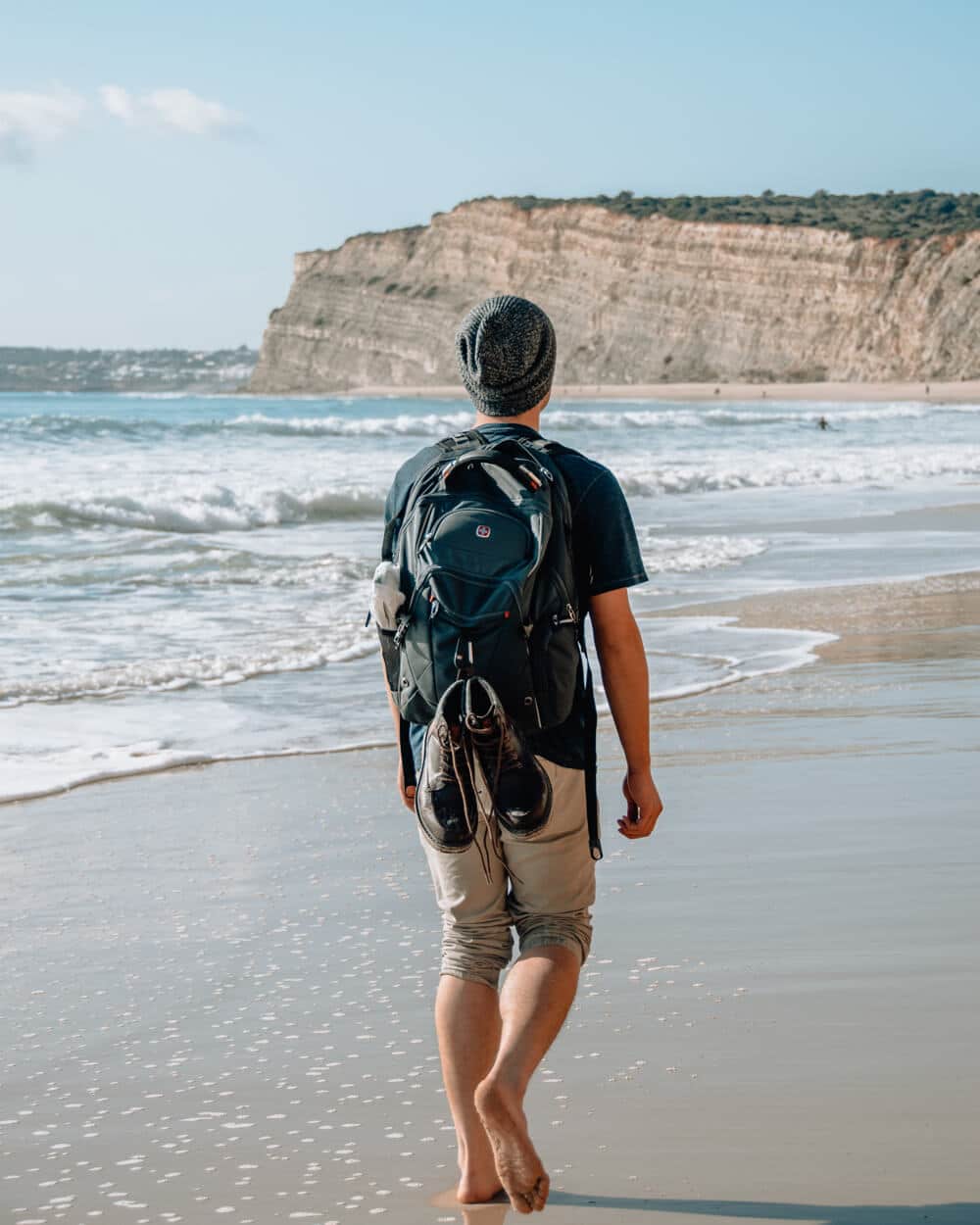 Person with a large backpack walking barefoot along a sandy beach toward the ocean, with cliffs in the distance.