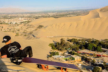 A tourist who has spent the morning sandboarding in Huacachina takes a break to sit on the high dune overlooking the spectacular view of the desert oasis below.