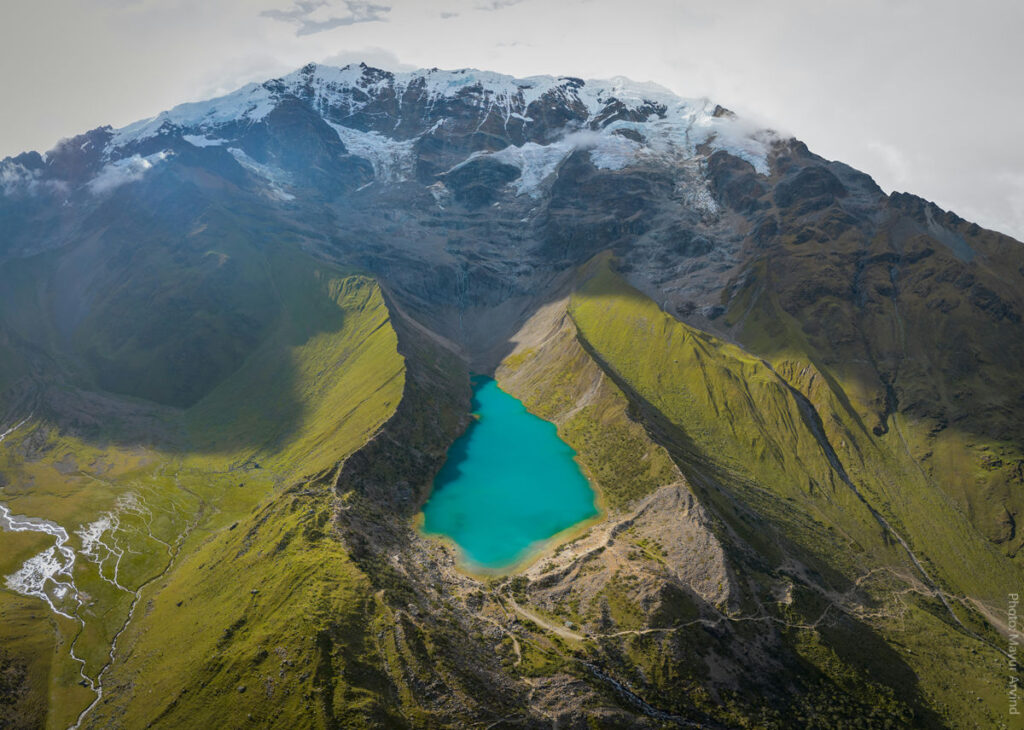 Aerial View of Humantay Lake — Emerald Glacial Lake on the Salkantay Trek, Cusco Peru | Peru Travel Blog Breathtaking aerial drone view of Humantay Lake, a vivid turquoise glacial lake nestled in a high Andean cirque below the snow-capped glaciers of Mount Humantay, surrounded by steep green mountain ridges and golden puna grasslands on the Salkantay Trek route, Cusco, Peru. Photo: Mayur Arvind.