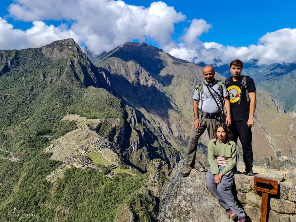Family at the Summit of Huayna Picchu — Machu Picchu with Kids, Cusco Peru | Peru Travel Blog A father and two children — a teenage boy and a young girl — posing at the summit of Huayna Picchu mountain, with the full aerial panorama of Machu Picchu's terraces and stone structures visible far below in the Urubamba canyon, Cusco, Peru.