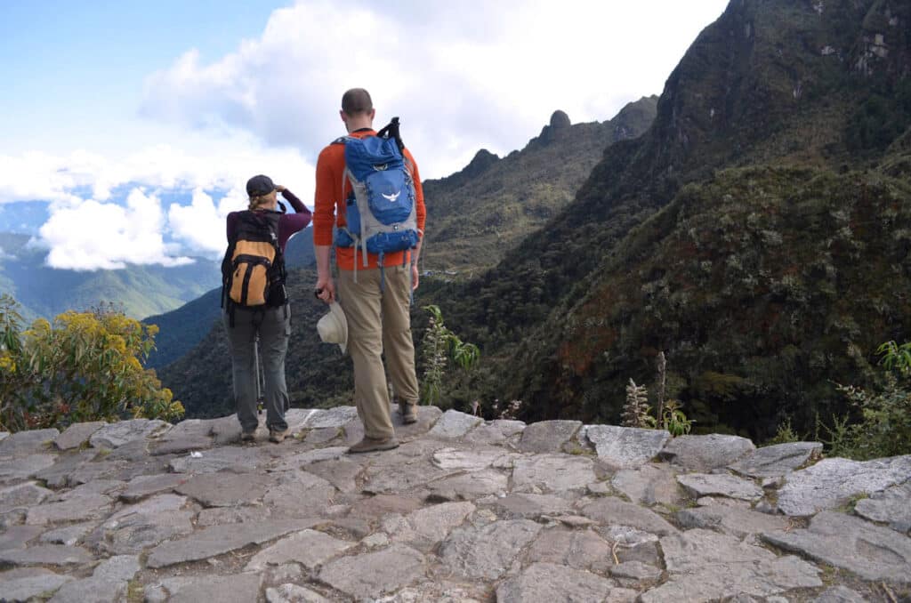 Two Hikers Pausing to Take in the View on the Inca Trail — Cusco, Peru | Peru Travel Blog Two hikers with backpacks standing on an ancient Inca stone-paved trail, pausing to take in the sweeping panorama of cloud-draped Andean mountain valleys and cloud forest on the classic 4-day Inca Trail to Machu Picchu, Cusco, Peru.