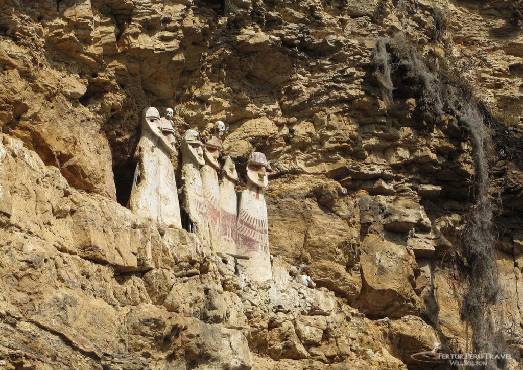 Karajía Sarcophagi — Ancient Chachapoya Cliff Funerary Figures, Amazonas Peru | Peru Travel Blog Six imposing Chachapoya clay sarcophagi set into a sheer limestone cliff face at Karajía in the Amazonas region of northern Peru, showing their elongated anthropomorphic bodies with painted red and white geometric designs, distinct facial features, and human skulls placed on top of several figures. Photo: Fertur Peru Travel / Will Skelton.