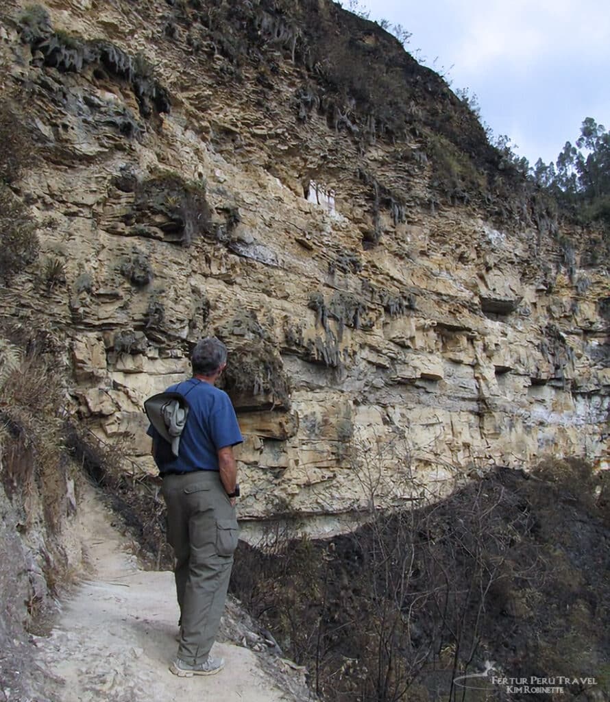 Approaching the Karajía Sarcophagi — Hiking the Cliff Trail, Amazonas Peru | Peru Travel Blog A Fertur Peru client standing on the narrow approach trail beside a towering limestone cliff face on the hike to the Karajía sarcophagi site in the Utcubamba Valley, Amazonas region of northern Peru, conveying the dramatic scale of the canyon walls. Photo: Fertur Peru Travel / Kim Robinette.
