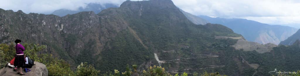 Machu Picchu Viewed from the Sun Gate (Inti Punku) — Inca Trail Finale, Peru | Peru Travel Blog Panoramic view from the Sun Gate (Inti Punku) on the Inca Trail, showing the Andean mountain landscape, the winding Hiram Bingham road, and the distant terraces of Machu Picchu visible on the right, with a traditionally dressed Andean woman seated on a rock in the foreground, Peru. Photo: Fertur Peru Travel.