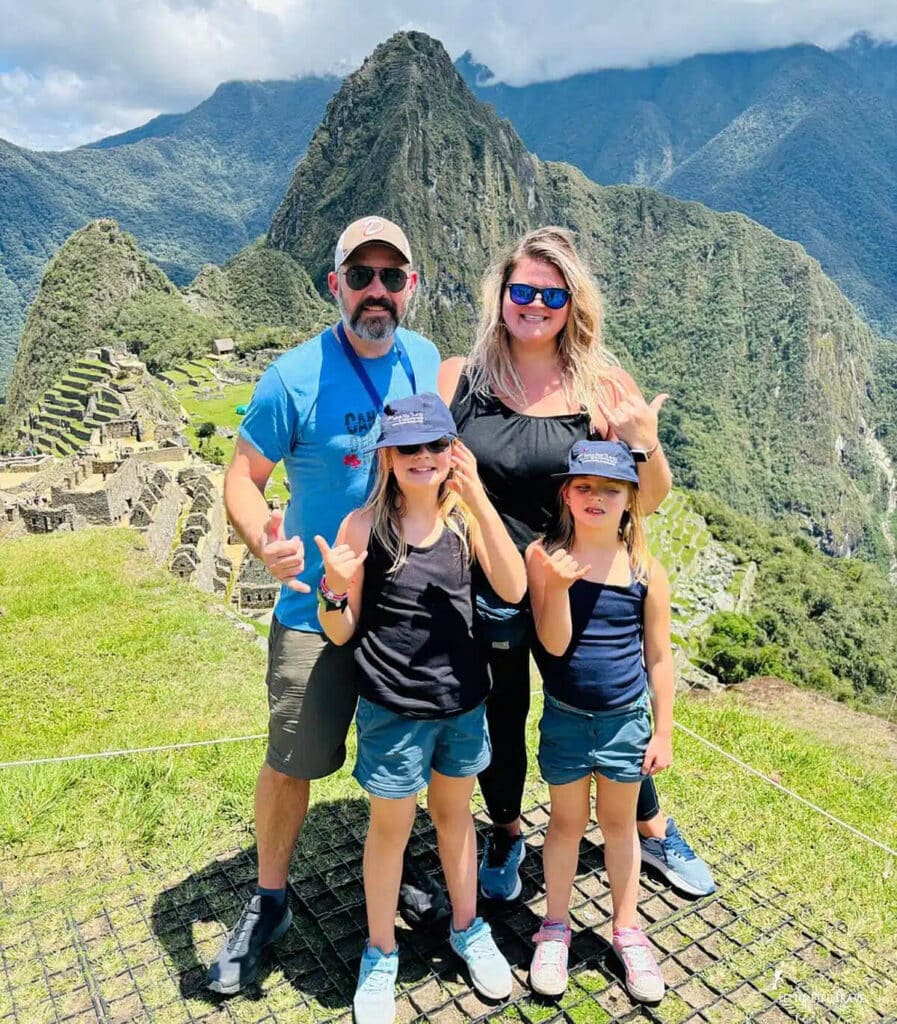 Family with Young Children at Machu Picchu — Peru Family Travel, Cusco | Peru Travel Blog A happy family of four — parents and two young girls wearing Fertur Peru Travel caps — giving shaka signs at the classic Machu Picchu viewpoint, with the iconic Inca citadel terraces, stone ruins, and Huayna Picchu peak visible behind them, Cusco, Peru.