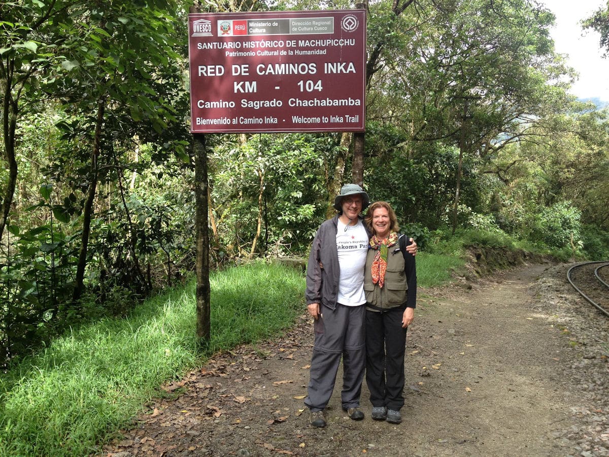 Inca Trail Trailhead at Km 104 — Camino Sagrado Chachabamba, Machu Picchu Peru | Peru Travel Blog A smiling couple posing under the official Red de Caminos Inka — Km 104 Camino Sagrado Chachabamba sign at the entrance to the 2-day Inca Trail within the Machu Picchu Historical Sanctuary, a UNESCO World Heritage Site, surrounded by lush cloud forest vegetation and a railway track, Cusco, Peru.