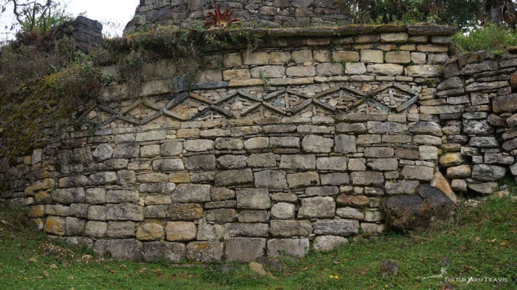 Tour Peru and visit the Chachapoya ruins of Kuelap | Peru Travel Blog Close-up of a Kuelap fortress stone wall displaying the distinctive Chachapoya decorative frieze of repeated diamond-shaped geometric motifs carved in stone, with moss, cloud forest vegetation, and a bromeliad plant growing along the top of the ancient masonry, Amazonas, Peru. Photo: Fertur Peru Travel.