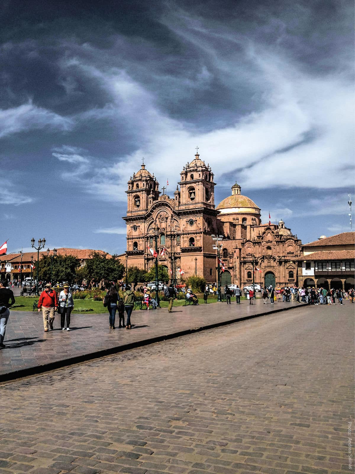 Plaza de Armas of Cusco — Jesuit Church of La Compañía de Jesús, Peru | Peru Travel Blog View of the Plaza de Armas of Cusco, Peru, dominated by the ornate baroque facade and twin bell towers of the Iglesia de La Compañía de Jesús, with its gilded dome, Peruvian flags, colonial arcaded buildings, and visitors strolling across the historic cobblestone square under a dramatic stormy sky. Photo: Andrea Salinas.