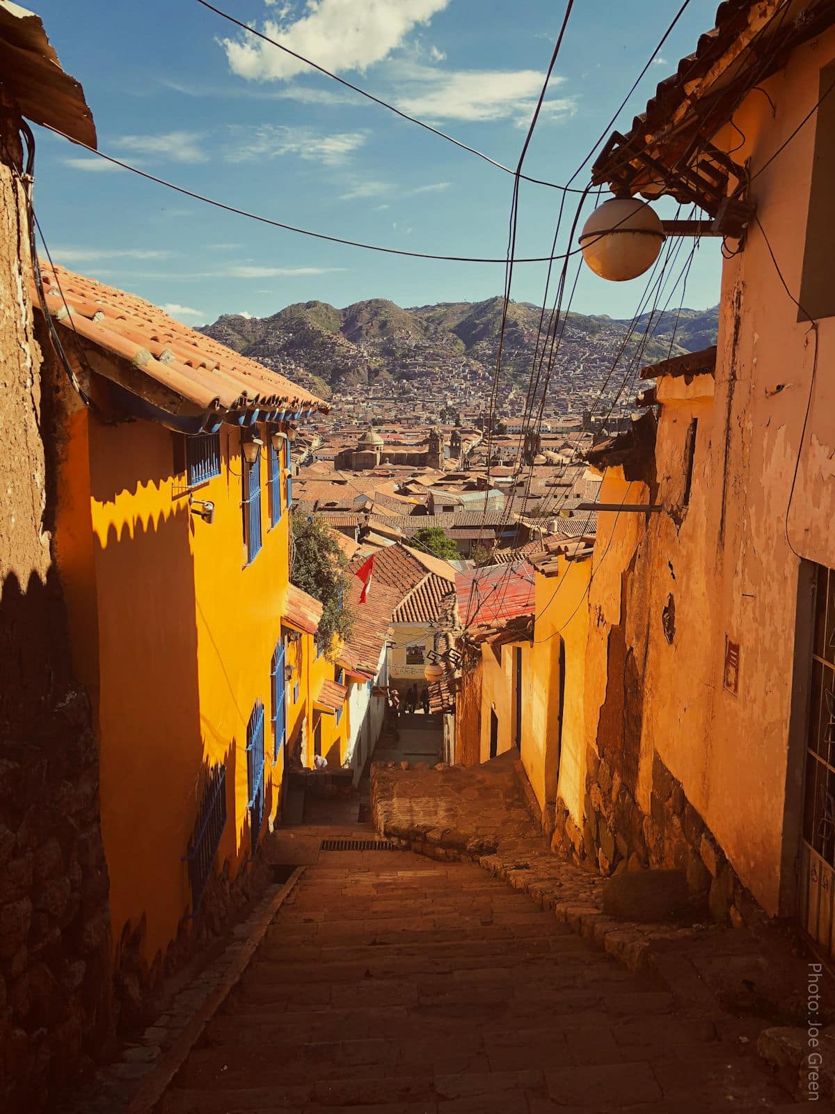 San Blas Neighborhood Cobblestone Street — Historic Cusco, Peru | Peru Travel Blog Steep cobblestone stairway descending through the San Blas neighborhood of Cusco, Peru, flanked by vibrant ochre and yellow colonial walls with blue wooden shutters, opening to a panoramic view of the city's terracotta rooftops and the surrounding Andean hills. Photo: Joe Green.