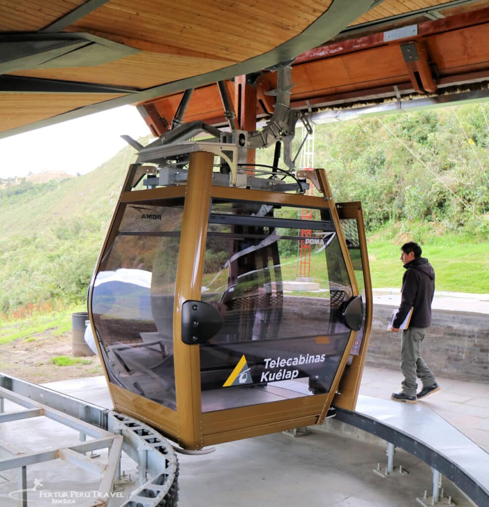 Telecabinas Kuélap Cable Car — Getting to Kuelap Fortress, Amazonas Peru | Peru Travel Blog A Telecabinas Kuélap cable car gondola at its boarding station in the Amazonas region of northern Peru, with the POMA-manufactured cabin bearing the official Telecabinas Kuélap branding, a staff member standing alongside, and cloud forest hills visible in the background. Photo: Fertur Peru Travel / Benjamin Ilka.