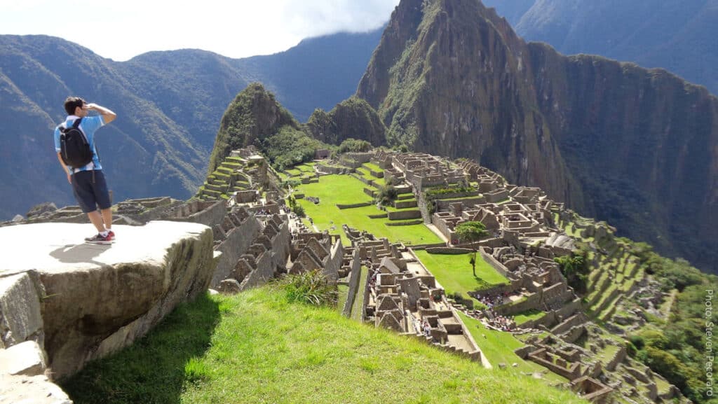 Machu Picchu Outer Circuit Viewpoint — Panoramic View of the Inca Citadel | Peru Travel Blog A visitor standing on a rocky outcrop on Machu Picchu's outer circuit gazes out over the full panorama of the Inca citadel, with its terraces, stone temples, and green plazas spread below, and the peak of Huayna Picchu rising dramatically through the clouds in the background. Photo: Steven Pecoraro.