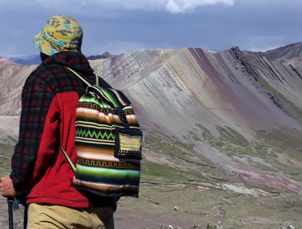 Visitor Gazing at Palcoyo Rainbow Mountain — High Andes, Cusco Peru | Peru Travel Blog A visitor wearing a colorful Andean woven backpack and bucket hat stands with a trekking pole, gazing at the vivid multicolored mineral-striped slopes of Palcoyo Rainbow Mountain in the high Andes of the Cusco region, Peru. Photo: Fertur Peru Travel.