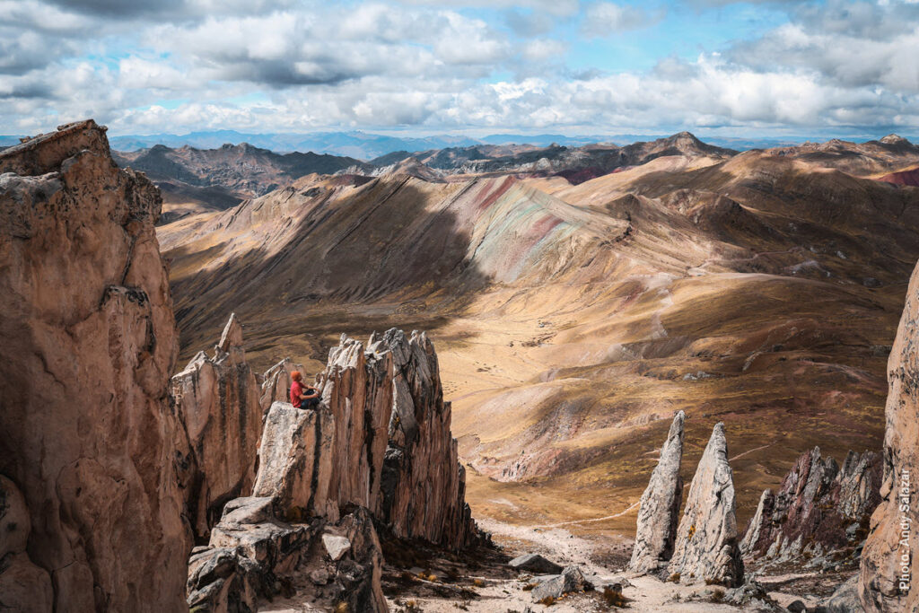 Palcoyo Rainbow Mountain from the White Stone Forest — Cusco, Peru | Peru Travel Blog A lone visitor seated on dramatic white quartzite rock spires of the Bosque de Piedras Blancas (White Stone Forest), with the sweeping high Andean plateau, the multicolored slopes of Palcoyo Rainbow Mountain, and endless mountain ranges stretching to the horizon under a dramatic cloudy sky, Cusco, Peru. Photo: Andy Salazar.