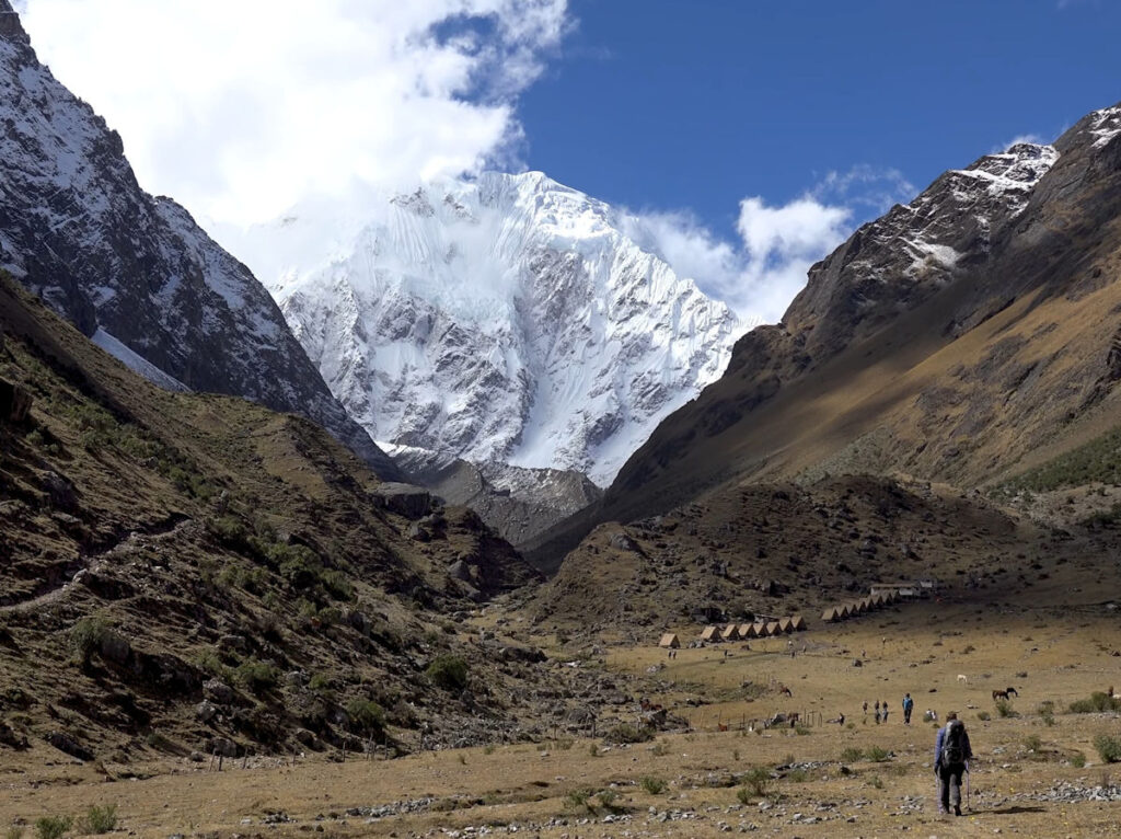 Salkantay Trek — Approaching the Glacier Base Camp, Cusco Peru | Peru Travel Blog A lone hiker with a backpack approaching the Salkantay trek base camp, a row of small tents nestled in the high Andean valley beneath the towering snow-covered glacier face of Mount Salkantay (6,271 m), with grazing horses and other trekkers visible in the golden highland meadow, Cusco, Peru.