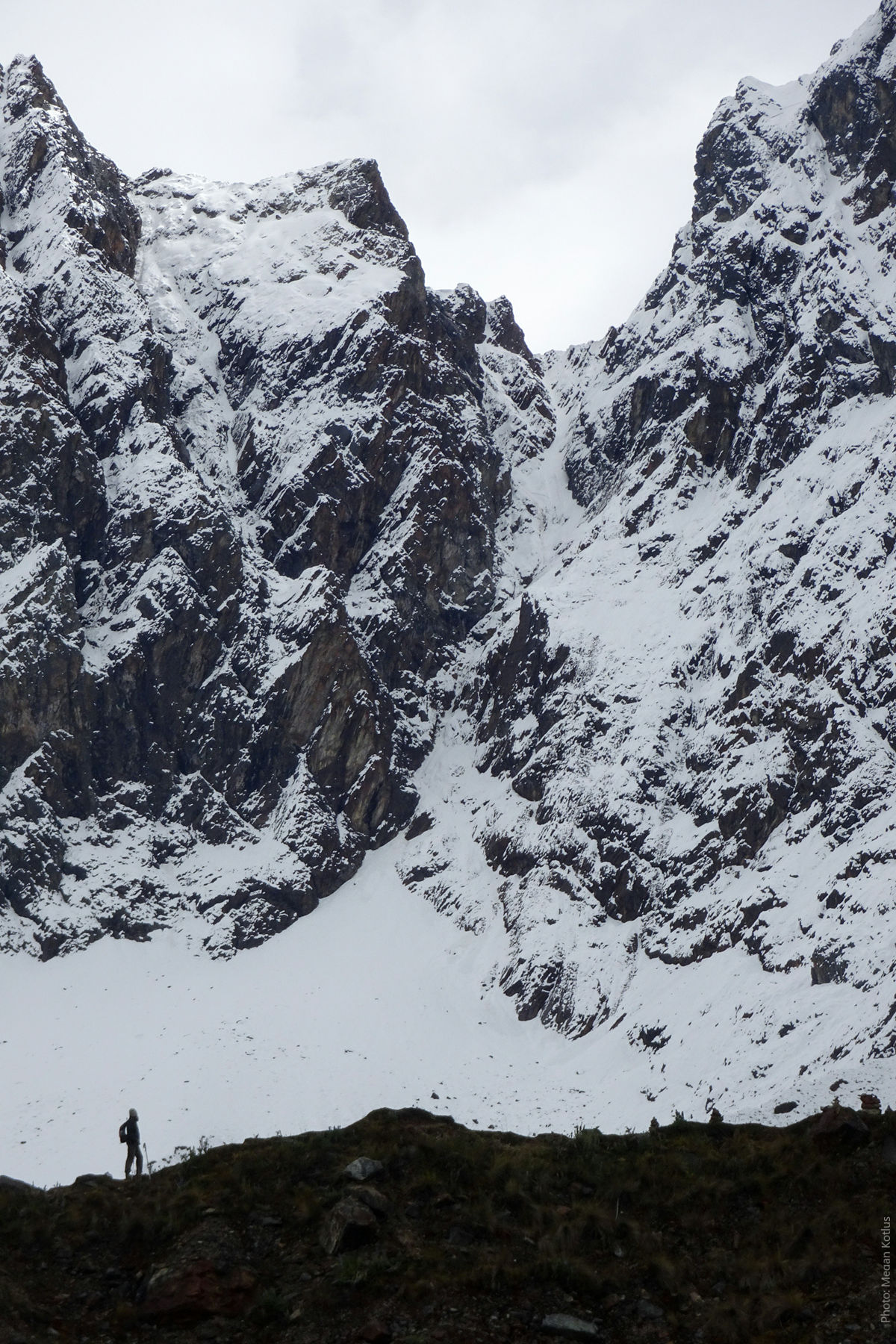 Lone Hiker Dwarfed by Salkantay Glacier Face — High Andes, Cusco Peru | Peru Travel Blog A lone silhouetted hiker with a trekking pole standing at the base of the massive snow and ice-covered rock face of Mount Salkantay, dramatically dwarfed by the towering glaciated cliffs rising hundreds of meters above, on the Salkantay Trek, Cusco, Peru. Photo: Megan Kotlus.