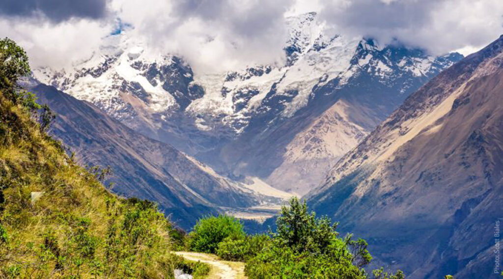 Salkantay Trek Valley View — Snow-Capped Andean Peaks and River Valley, Cusco Peru | Peru Travel Blog Sweeping panoramic view of a deep glacial valley on the Salkantay Trek, with snow-capped Andean peaks emerging through dramatic storm clouds, a river valley floor visible far below, and lush cloud forest vegetation in the foreground, Cusco, Peru. Photo: Scott Biales.