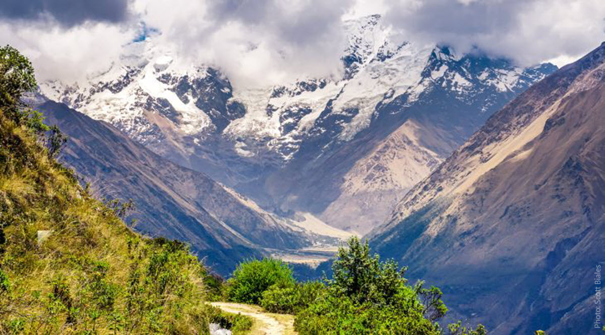 Salkantay Trek Valley View — Snow-Capped Andean Peaks and River Valley, Cusco Peru | Peru Travel Blog Sweeping panoramic view of a deep glacial valley on the Salkantay Trek, with snow-capped Andean peaks emerging through dramatic storm clouds, a river valley floor visible far below, and lush cloud forest vegetation in the foreground, Cusco, Peru. Photo: Scott Biales.