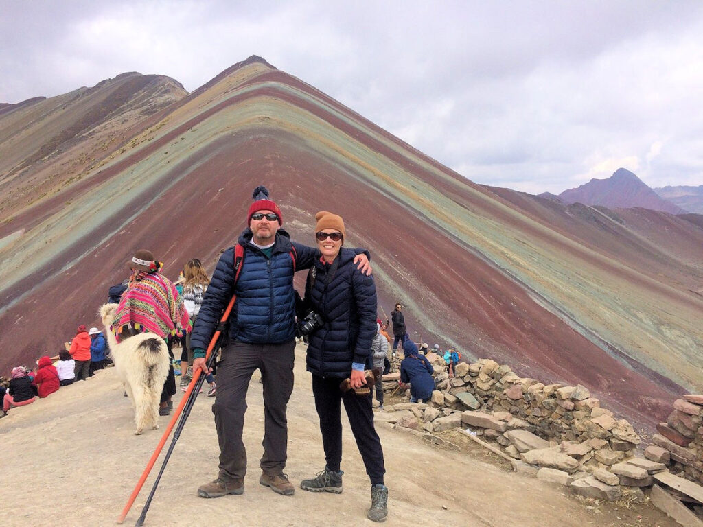 Couple at the Summit of Vinicunca Rainbow Mountain — Cusco, Peru | Peru Travel Blog A couple in warm winter gear with trekking poles posing at the summit of Vinicunca Rainbow Mountain, with the iconic pyramidal multicolored slopes of red, purple, yellow, and green clearly visible behind them, alongside other visitors, a llama, and a local guide in traditional dress, Cusco, Peru.