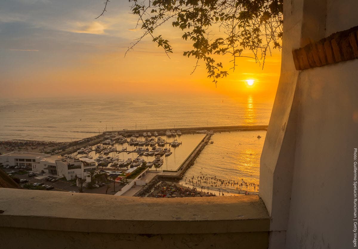 Sunset Over the Marina and Pacific Coast Viewed from the Barranco Clifftop | Lima, Peru | Peru Travel Blog A spectacular clifftop sunset view over Lima's coastline, framed through a white colonial archway in the Barranco district. Photo: Guillermo Spelucin Runciman / Getty Images via Canva Pro.