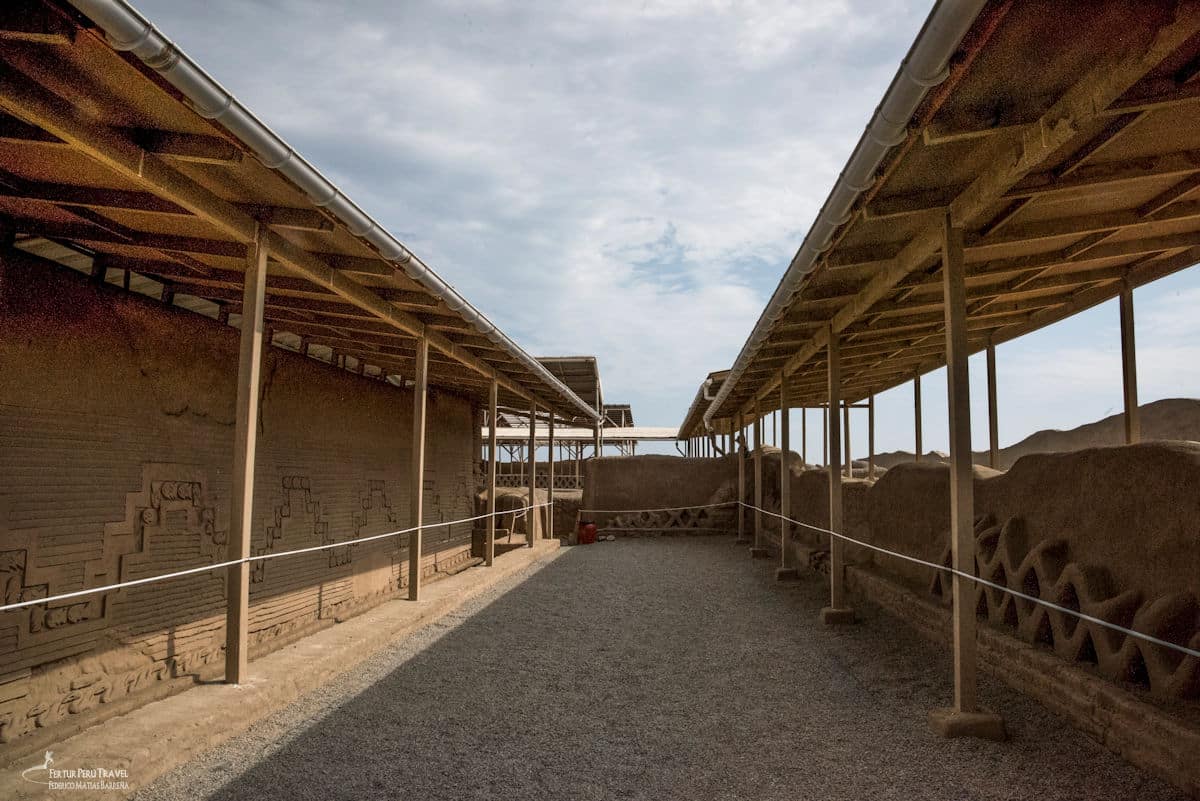 Walking the Visitor Corridor at Chan Chan | Chimú Adobe Friezes | Trujillo Peru | Peru Travel Blog A gravel visitor walkway running between two rows of protective metal roofing structures at Chan Chan, with elaborately carved adobe walls displaying Chimú stepped geometric and wave-pattern friezes on both sides, Trujillo, La Libertad, Peru. Photo: Fertur Peru Travel / Federico Matías Barreña.