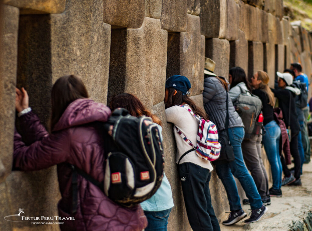 Cusco Sacred Valley Tour - Ollantaytambo | Peru Travel Blog Tourists with backpacks lean against a tall stone wall, peering through its gaps during a guided visit.