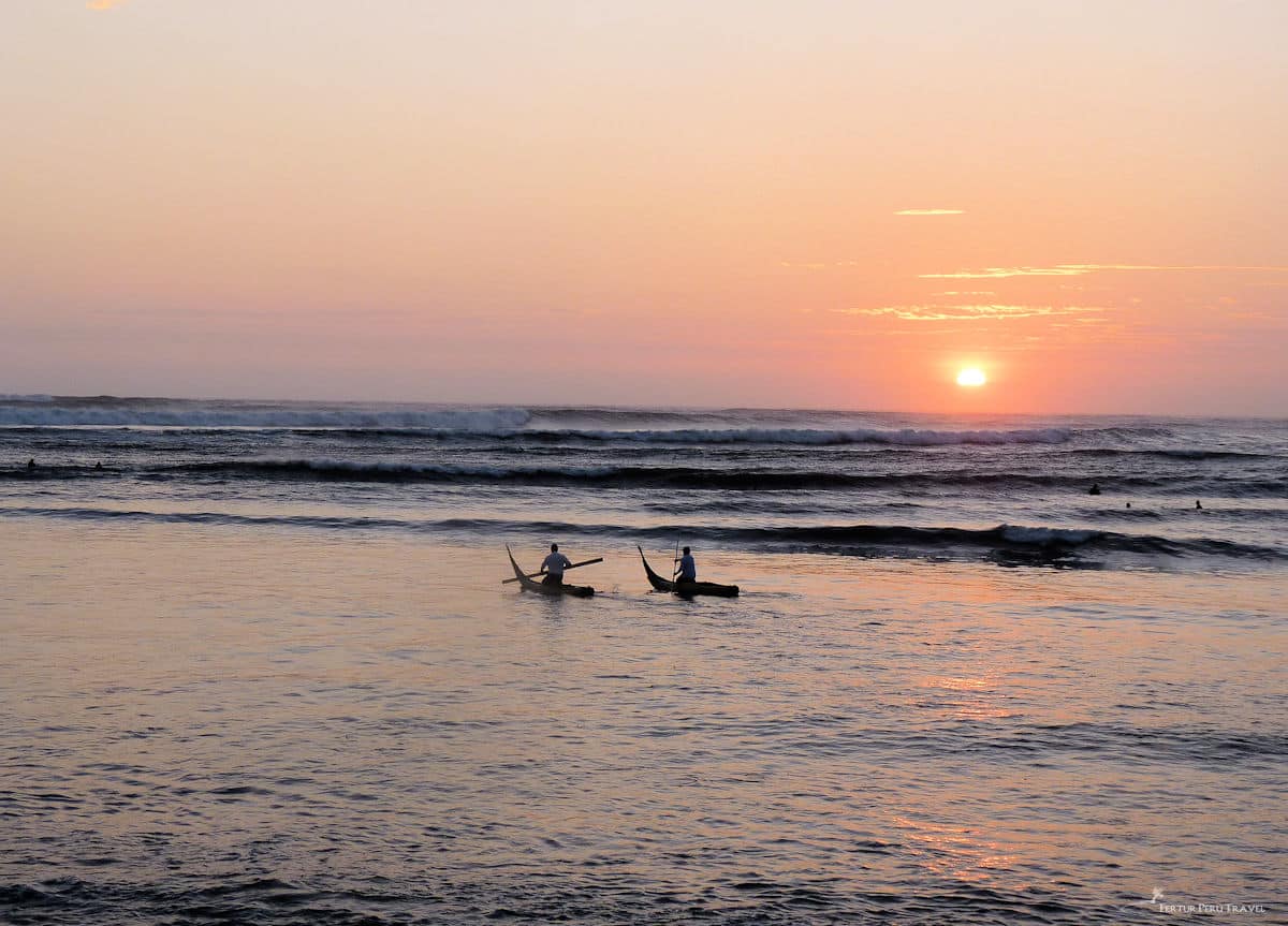 Huanchaco Fishermen on Caballitos de Totora at Sunset | Peru Travel Blog Two fishermen paddling traditional caballito de totora reed boats through the surf at sunset on the beach of Huanchaco, with rolling Pacific waves and a vivid orange and pink sky reflected on the water, Trujillo, La Libertad, Peru. Photo: Fertur Peru Travel.