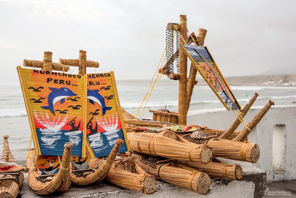 Handcrafted Caballito de Totora Reed Boat Souvenirs at Huanchaco, Peru | Peru Travel Blog Miniature totora reed boat souvenirs with painted sails reading "Huanchaco Peru," displayed on a wall with the ocean in the background