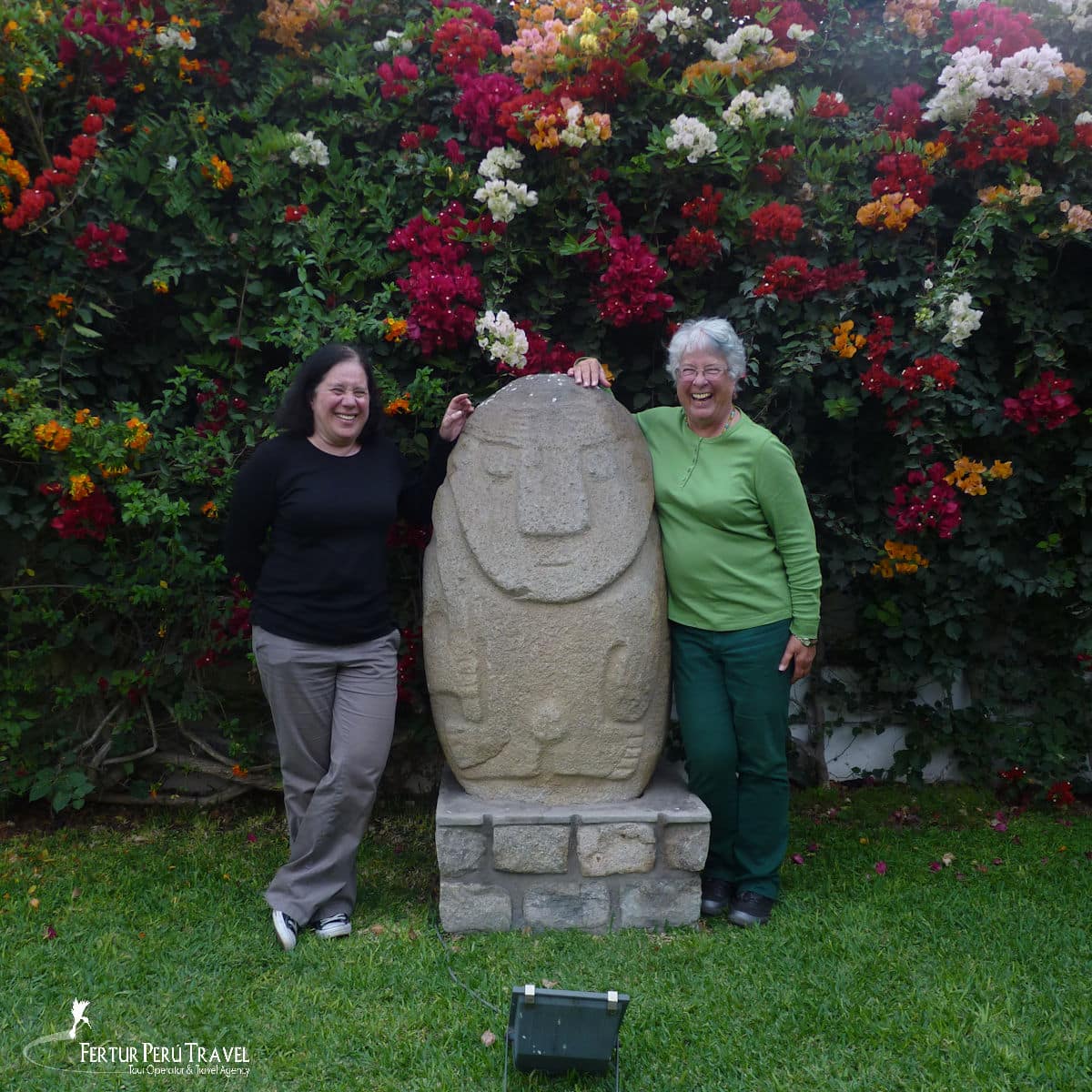 Two Travelers Posing with a Pre-Columbian Stone Idol in the Garden of the Larco Museum, Lima, Peru | Peru Travel Blog Two Fertur Peru Travel clients share a laugh beside a large pre-Columbian stone idol — likely of Recuay or Huari origin — displayed in the legendary garden of the Museo Larco in the Pueblo Libre district of Lima. The garden's spectacular explosion of multicolored bougainvillea in full bloom — red, white, orange, and pink — provides a vivid backdrop for one of Lima's most beloved and photogenic museum experiences.