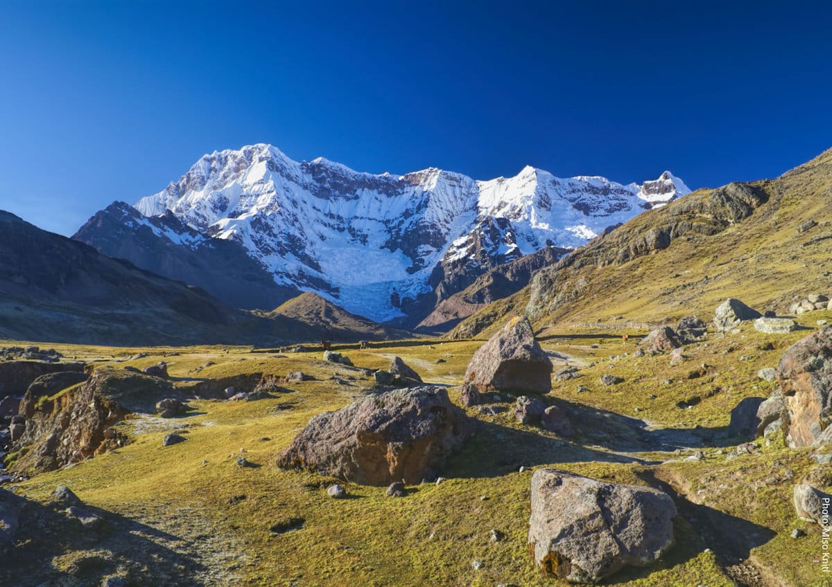 Mount Ausangate Glacier and High-Altitude Puna Landscape, Ausangate Circuit, Cusco, Perund High-Altitude Puna Landscape - Ausangate Circuit | Peru Travel Blog The snow-capped and glaciated summit of Mount Ausangate rising above a high-altitude puna grassland scattered with boulders under a deep blue Andean sky, Cusco region, Peru