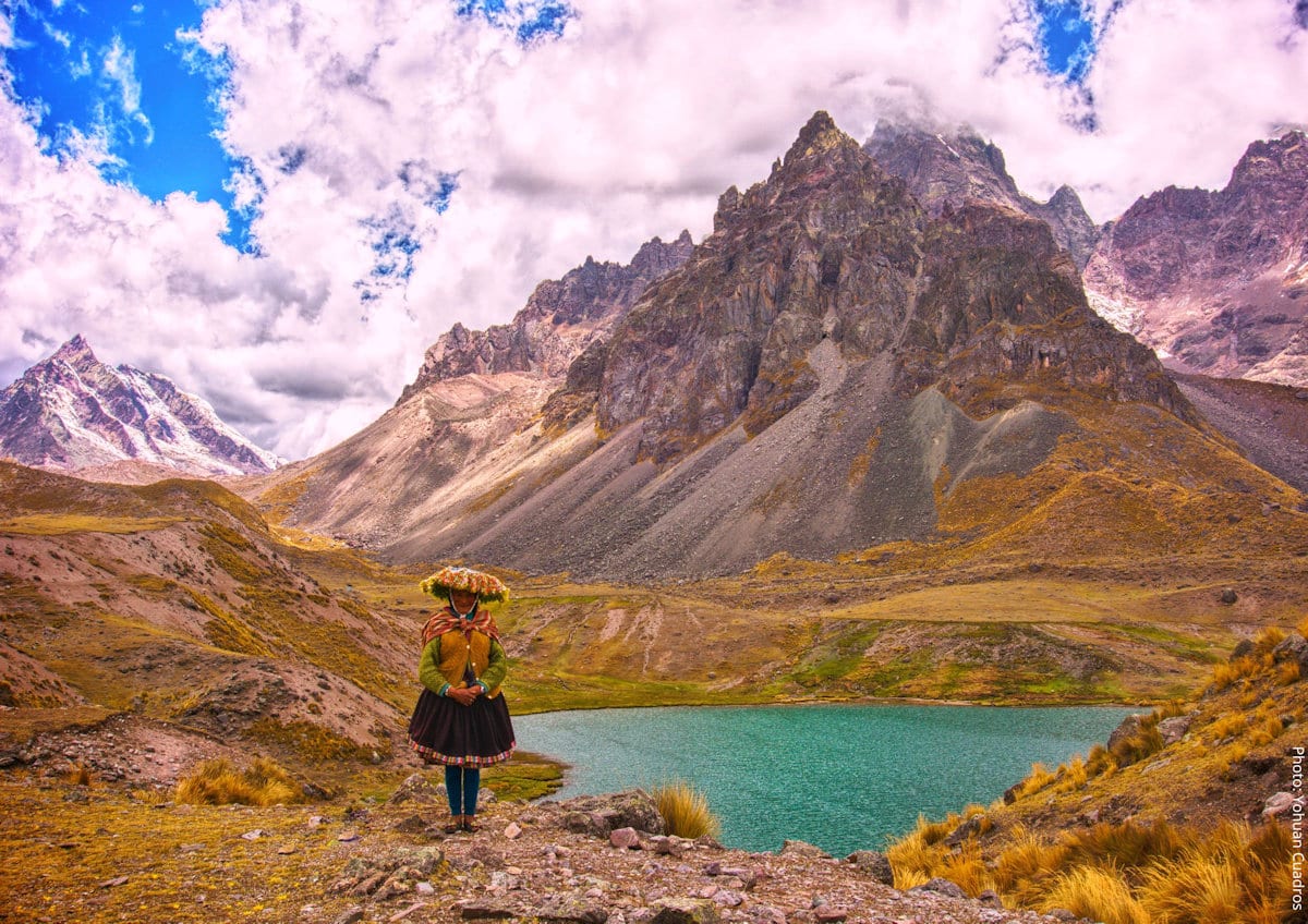Quechua Woman in Traditional Dress Before Mount Ausangate and a Glacial Lake, Cusco, Peru | Peru Travel Blog A Quechua woman in traditional Andean dress and a flower crown stands before a turquoise glacial lake with the jagged snow-capped peaks of Mount Ausangate rising dramatically behind her, Cusco region, Peru