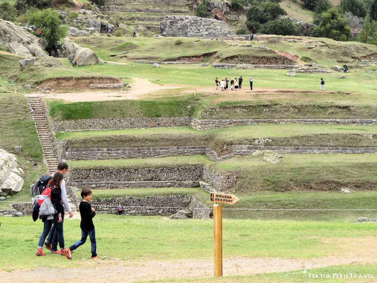 Sacsayhuaman Cusco - Family Trip in October | Peru Travel Blog A family with two children walking along the green terraced grounds of the Sacsayhuaman Inca fortress above Cusco, with a wooden Mirador/Viewpoint directional sign in the foreground, stone terraces, and other visitors visible across the expansive archaeological site, Cusco, Peru. Photo: Fertur Peru Travel.