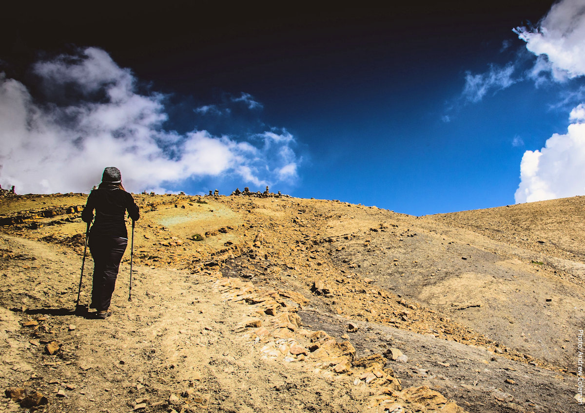 Solo Ausangate Trekker Climbing the Final Ascent to Vinicunca Rainbow Mountain | Peru Travel Blog A lone trekker with a walking pole climbs a steep rocky trail toward a ridge lined with stone cairns under a dramatic deep blue sky, on the final ascent to Vinicunca Rainbow Mountain, Cusco region, Peru