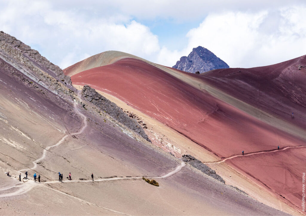 Trekkers on the Trail Through Red Valley Near Vinicunca Rainbow Mountain, Cusco, Peru | Peru Travel Blog A winding trail cuts through the dramatically colored red and ochre slopes of Red Valley near Vinicunca Rainbow Mountain, with a group of trekkers dwarfed by the vast mineral-streaked landscape and a rocky peak rising in the background, Cusco region, Peru