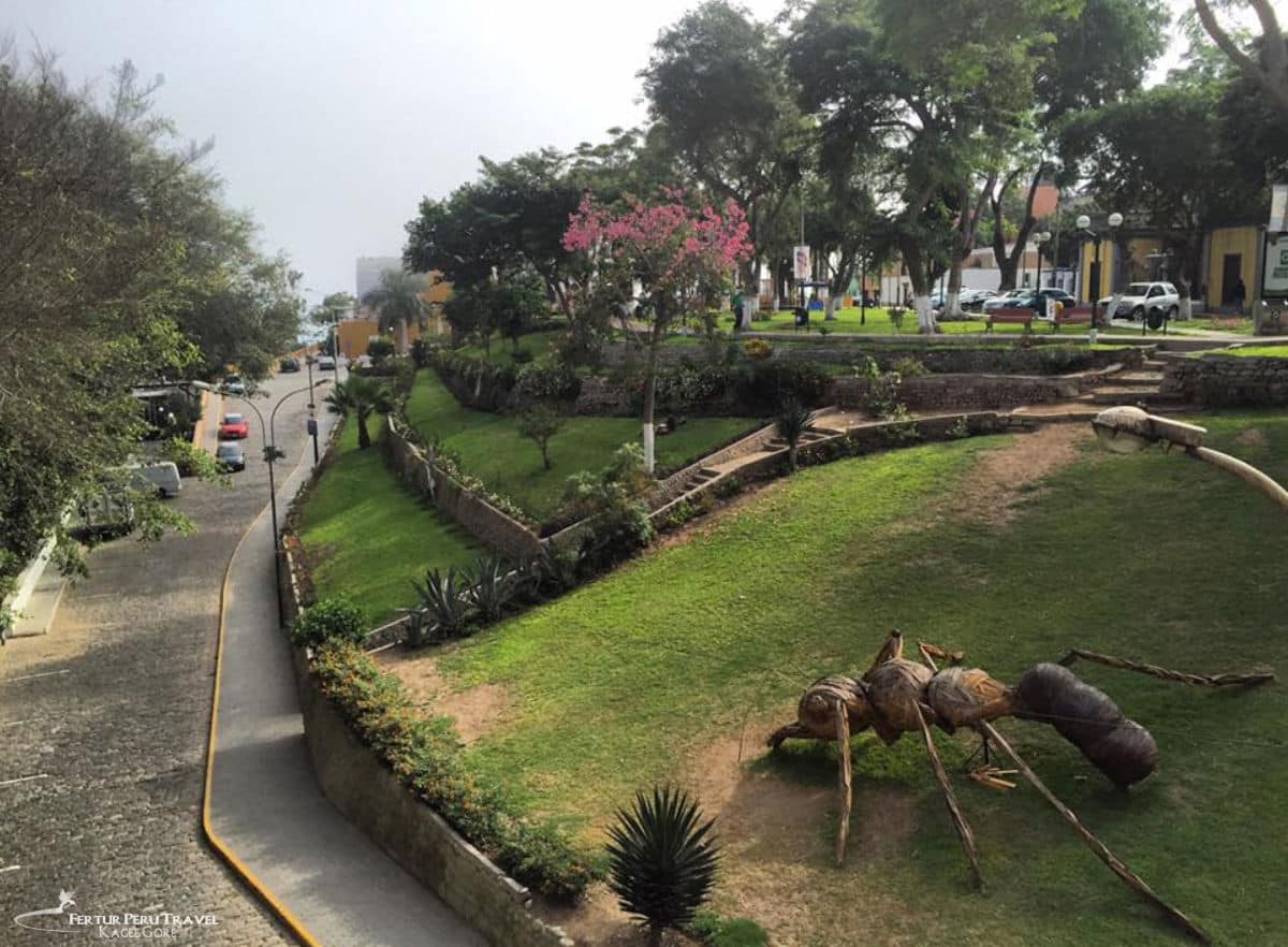 Giant Ant Sculpture in the Terraced Gardens of Parque Municipal, Barranco | Lima, Peru | Peru Travel Blog A large sculptural giant ant made from natural wood and branches installed on the terraced green lawns of Parque Municipal in the bohemian Barranco district of Lima, Peru, with a flowering pink tree and the district's streets visible beyond.