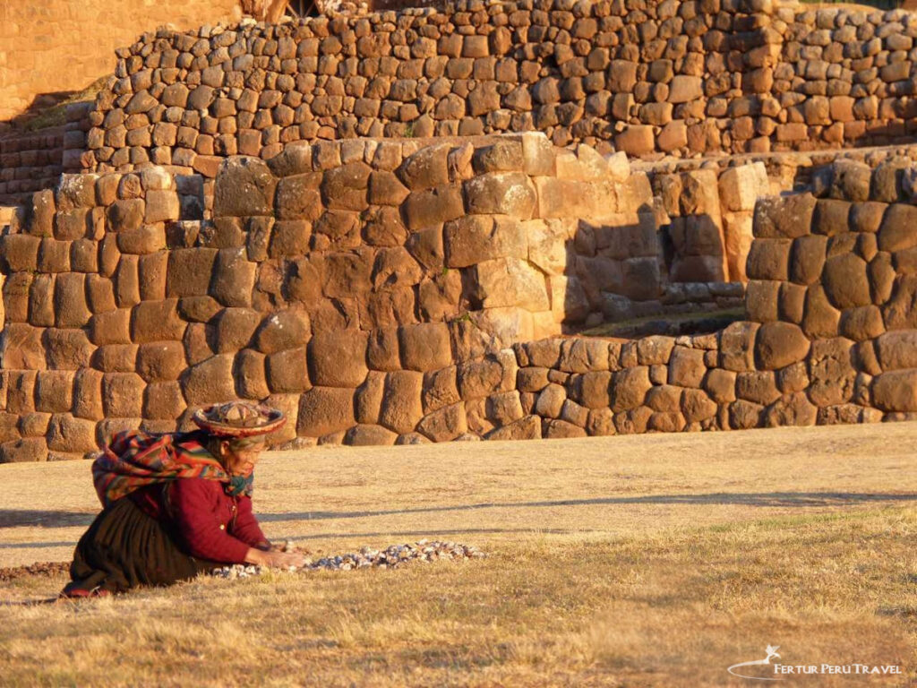 Quechua Woman Processing Chuño at Chinchero, Sacred Valley | Cusco, Peru | Peru Travel Blog A Quechua woman in traditional dress and a wide-brimmed hat kneels on dry golden grass sorting chuño freeze-dried potatoes before the ancient Inca stone walls of Chinchero in warm late-afternoon light, Sacred Valley, Cusco, Peru