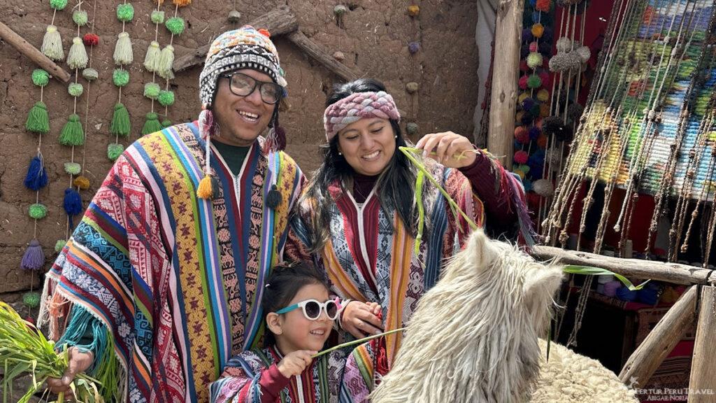 Family in Traditional Andean Ponchos Feeding an Alpaca at a Weaving Community, Sacred Valley, Cusco, Peru | Peru Travel Blog A smiling family of three — father, mother, and young daughter — dressed in colorful traditional Andean ponchos and knit hats feed grass to a fluffy alpaca at a weaving community, surrounded by hanging yarn tassels and textiles, Sacred Valley, Cusco, Peru