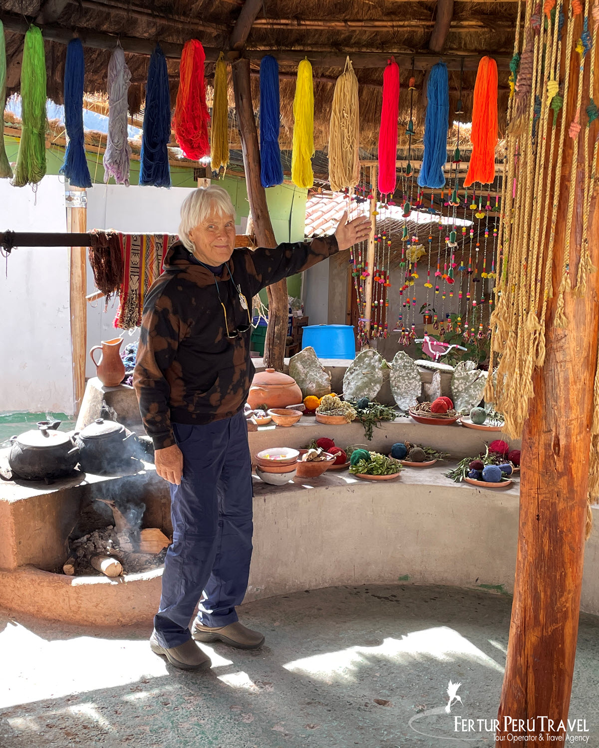 Traveler Exploring Traditional Natural Dye Materials at a Weaving Cooperative in Chinchero, Sacred Valley, Peru | Peru Travel Blog A Fertur Peru Travel client takes in a hands-on demonstration of traditional Andean natural dyeing at a weaving cooperative in Chinchero, high above the Sacred Valley at 3,762 meters. Brilliant skeins of alpaca and sheep wool — dyed with cochineal, indigo, muña, and other native plants — hang overhead in a full spectrum of colors, while clay pots, grinding stones, dried herbs, raw plant materials, and pre-dyed wool balls are arranged on a low curved adobe bench below.