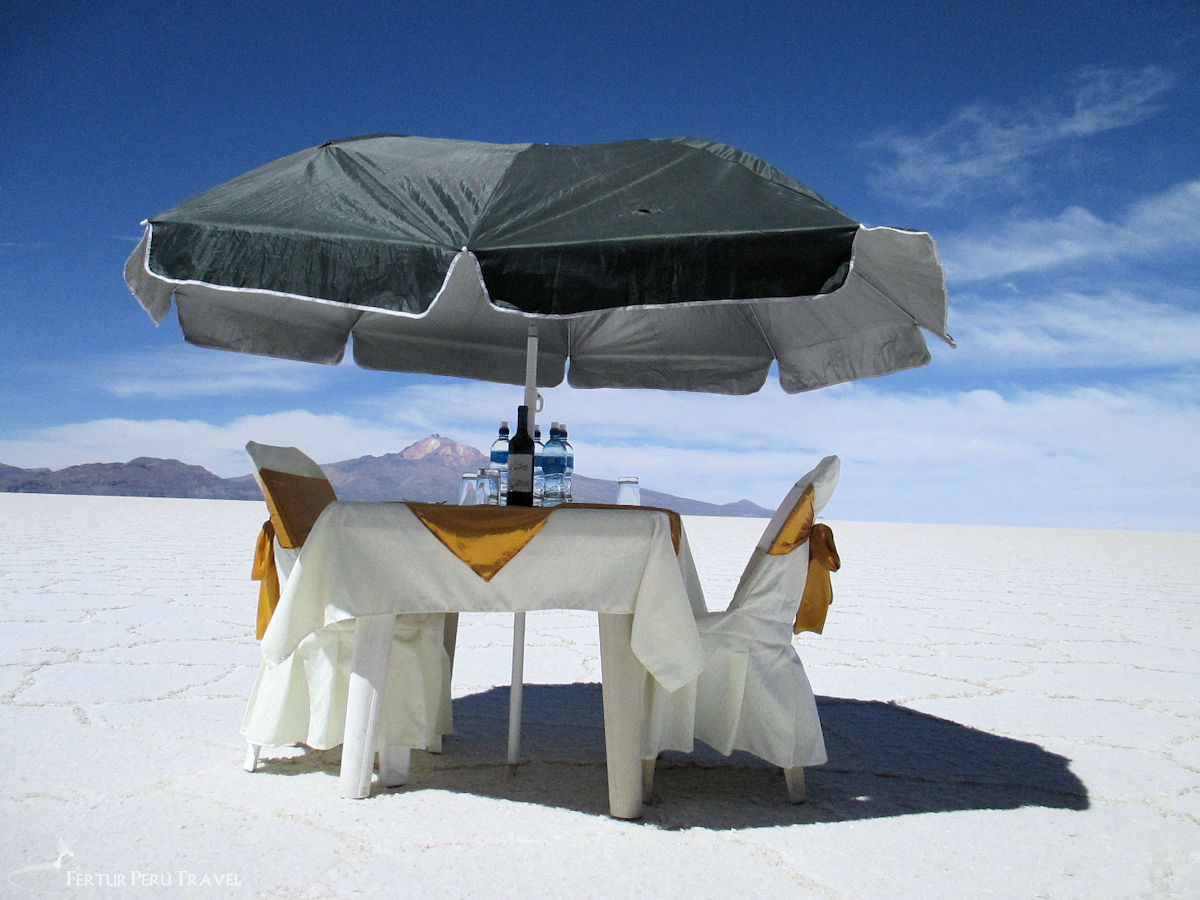 Private Lunch for Two Set Up on the Salar de Uyuni Salt Flats, Bolivia | Peru Travel Blog A white-clothed table for two with gold sash chairs, a wine bottle, and water set under a large green umbrella in the middle of the blindingly white Salar de Uyuni salt flats, with a volcanic peak rising in the distance, Bolivia