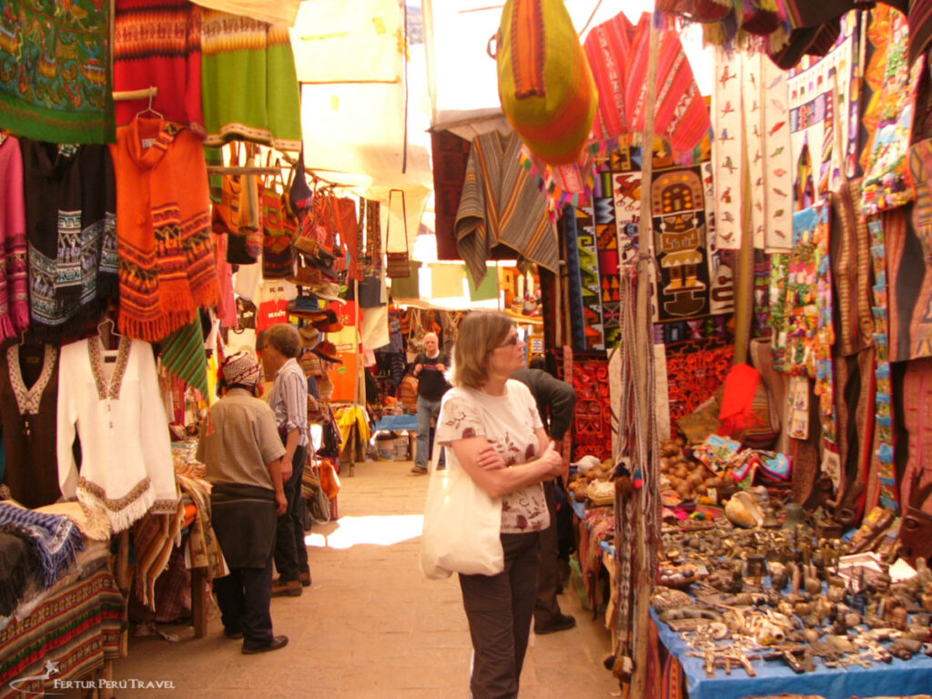 Tourists Browsing the Colorful Artisan Market at Pisac | Sacred Valley, Cusco, Peru | Peru Travel Blog Tourists browse a vibrant covered artisan market lane packed with hanging alpaca textiles, woven ponchos, tapestries, ceramics, and silver crafts at the Pisac market in the Sacred Valley, Cusco, Peru