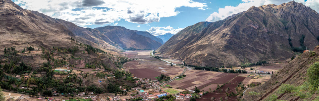 Panoramic View of the Sacred Valley of the Incas and Urubamba River, Cusco, Peru | Peru Travel Blog A sweeping panoramic view of the Sacred Valley of the Incas showing the Urubamba River winding through a broad valley floor of patchwork agricultural fields, with a small Andean town nestled at the base of dramatic steep mountain walls, Cusco, Peru