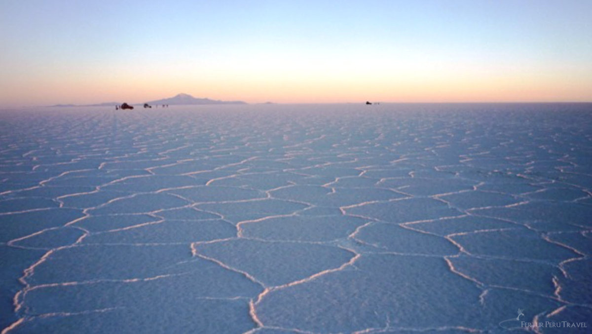 Hexagonal Salt Crust Patterns at Sunset on the Salar de Uyuni, Bolivia | Peru Travel Blog The geometric hexagonal salt crust patterns of the Salar de Uyuni stretch to the horizon at sunset, tinted in soft blue and peach tones, with distant 4x4 vehicles and a volcanic silhouette on the horizon, Bolivia