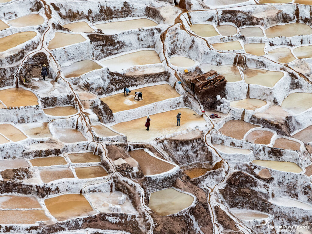 Salt Workers Harvesting at the Salineras de Maras Terraced Salt Ponds, Sacred Valley | Cusco, Peru | Peru Travel Blog Aerial close-up view of workers harvesting salt from the thousands of white-crusted terraced evaporation ponds of the Salineras de Maras, with golden brine-filled pools and salt sacks visible, Sacred Valley, Cusco, Peru.