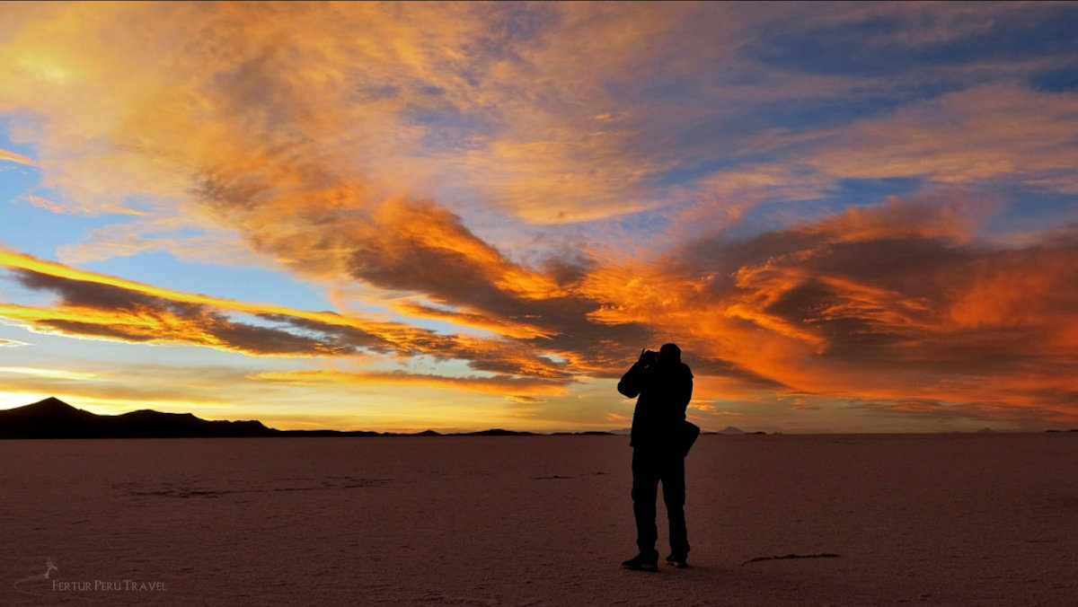 Photographer Silhouetted Against a Fiery Sunset Sky on the Salar de Uyuni Salt Flats, Bolivia | Peru Travel Blog The dramatic silhouette of a lone photographer shooting a blazing orange and gold sunset sky over the flat horizon of the Salar de Uyuni salt flats, with volcanic peaks visible in the distance, Bolivia