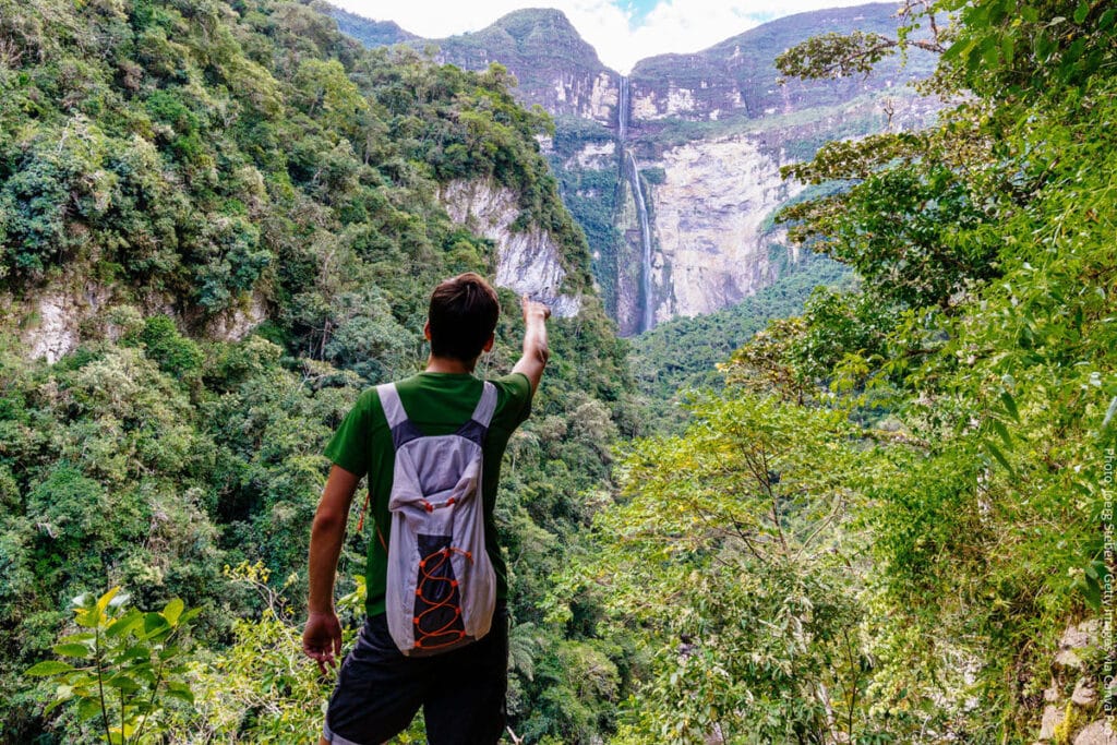 Trekker Points Toward Gocta Falls from the Cloud Forest Trail, Chachapoyas, Amazonas, Peru | Peru Travel Blog A male trekker with a daypack stands on the cloud forest trail pointing toward the full two-tiered cascade of Gocta Falls dropping down a sheer cliff face surrounded by lush green canyon walls, Amazonas region, Peru. Photo: Joerg Steber / Getty Images via Canva Pro.