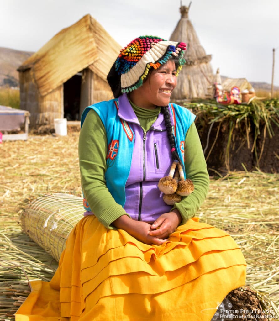 Uros Woman in Traditional Dress on the Floating Reed Islands of Lake Titicaca, Puno, Peru | Peru Travel Blog A smiling Uros woman in a vibrant golden yellow skirt, colorful embroidered vest, and a distinctive multicolored pompom hat sits on the totora reed surface of a floating island with traditional reed dwellings behind her, Lake Titicaca, Puno, Peru